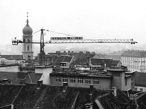 Schwarzweißfoto einer Stadtlandschaft mit einem Baukran mit der Aufschrift „KAISER ROTTLER“ über den Dächern. Im Hintergrund sind eine Kirchenkuppel und ein Turm vor einem bewölkten Himmel sichtbar.