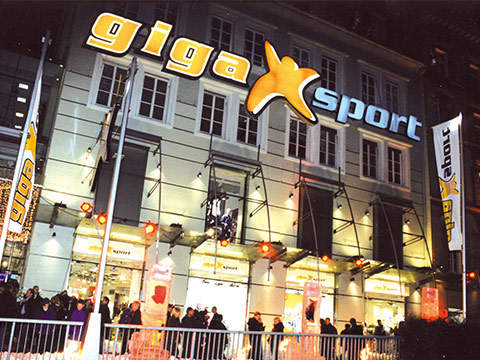 Nachtansicht eines großen, beleuchteten Ladenlokals mit einem auffälligen „Gigasport“-Schild. Das Gebäude hat mehrere Stockwerke und ist mit Lichtern dekoriert. Draußen hat sich eine Menschenmenge versammelt und Banner mit dem Gigasport-Logo sind zu sehen.