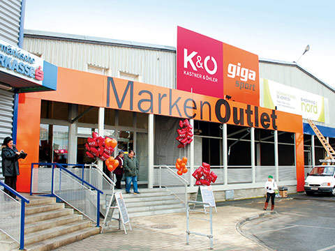 Eingang eines Marken Outlet-Geschäfts mit orange-schwarzer Beschilderung. An Ständern draußen hängen rote und orangefarbene Luftballons. Eine Person in einer schwarzen Jacke macht ein Foto, eine andere Person geht daneben. Eine Leiter lehnt am Gebäude.