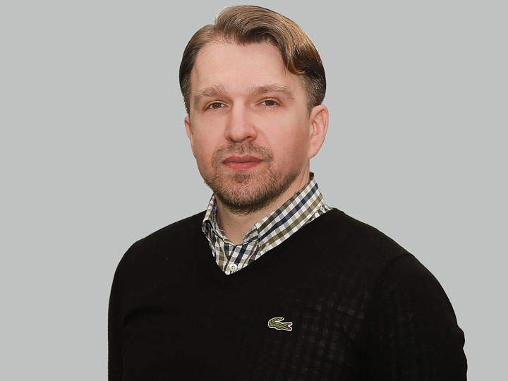 Ein Mann mit kurzen Haaren und Bart trägt einen schwarzen Pullover über einem karierten Hemd. Er steht vor einem schlichten grauen Hintergrund und blickt in die Kamera.