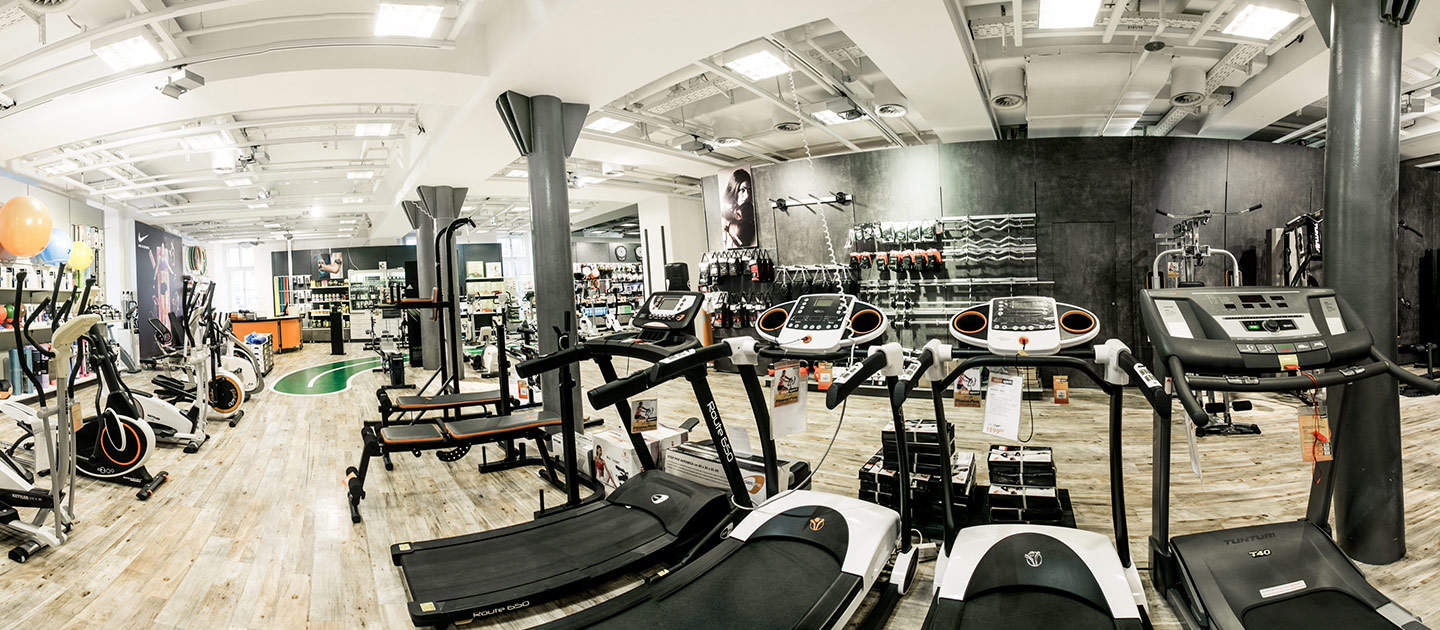 Ein geräumiger Fitnessgeräteladen mit Holzboden zeigt verschiedene Fitnessgeräte. Laufbänder säumen den Vordergrund, während im Hintergrund Heimtrainer und Zubehör zu sehen sind. Helle Deckenbeleuchtung erhellt den Raum.