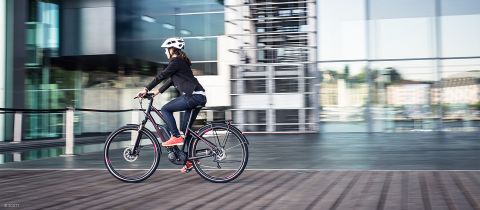 Eine Person mit Helm und Freizeitkleidung fährt mit einem Elektrofahrrad auf einem Holzweg. Im Hintergrund ist ein modernes Glasgebäude mit Spiegelungen und einer verschwommenen Stadtlandschaft zu sehen.