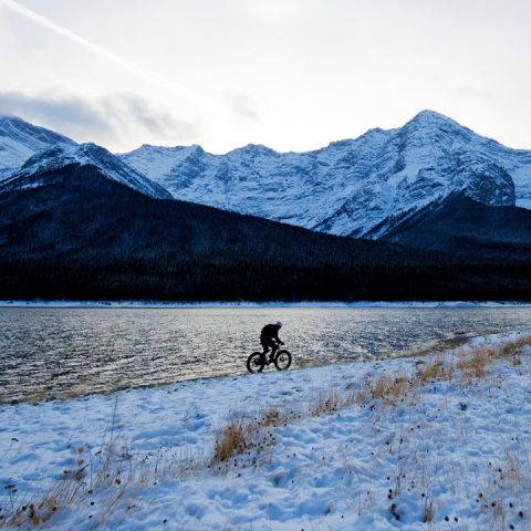 Silhouette einer Person, die auf einem verschneiten Weg neben einem See Fahrrad fährt. Im Hintergrund erheben sich schneebedeckte Berge unter einem bewölkten Himmel. Die Landschaft ist ruhig und die Berge spiegeln sich auf der Wasseroberfläche.
