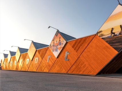 Modernes Gebäude mit sechseckiger Holzfassade und dem Schriftzug „ggysport“. Im oberen Bereich sind große Poster zu sehen, darunter Bilder von Menschen, die Outdoor-Aktivitäten nachgehen. Im Hintergrund ist ein strahlend blauer Himmel zu sehen.