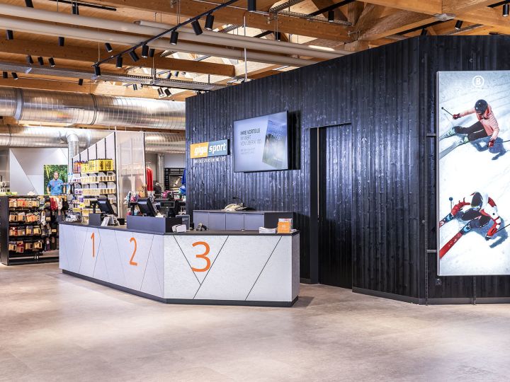 Ein modernes Interieur eines Sportgeschäfts mit einem eleganten Kundendienstschalter mit den Aufschriften 1, 2, 3. An der Wand dahinter sind Sportbilder zu sehen und es gibt ordentlich angeordnete Regale mit verschiedenen Sportartikeln. Die Decke weist freiliegende Balken und Rohrleitungen auf.