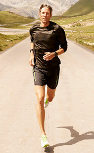 Eine Person joggt auf einer asphaltierten Straße, umgeben von grünen Hügeln und Bergen. Sie trägt ein dunkles Langarmshirt, schwarze Shorts und Laufschuhe. Die Sonne wirft einen Schatten auf die Straße.