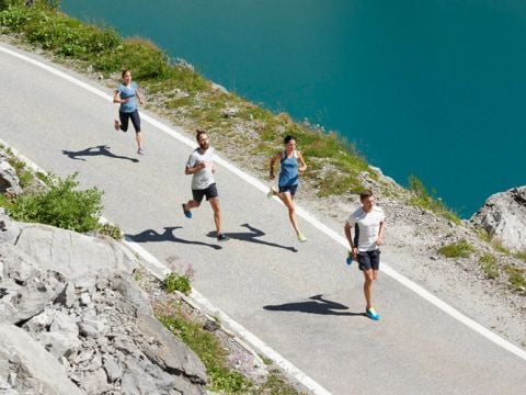Vier Personen joggen auf einem gepflasterten Weg neben einem klaren, blauen See. Die Szene ist von Felsen und Grün umgeben. Die Gruppe hat sich über die Straße verteilt und genießt die Bewegung im Freien.