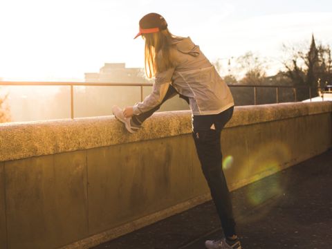 Eine Person in leichter Jacke und Mütze streckt bei Sonnenaufgang oder Sonnenuntergang ihr Bein auf einem Felsvorsprung aus. Sie befindet sich im Freien auf einem Pfad mit einer malerischen, sonnenbeschienenen Kulisse aus Bäumen und entfernten Gebäuden.