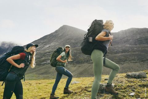 Drei Personen wandern einen grasbewachsenen Berghang hinauf. Sie tragen Wanderausrüstung und große Rucksäcke. Der Himmel ist bewölkt und im Hintergrund ist ein felsiger Berggipfel zu sehen.