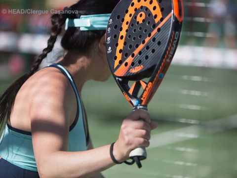 Eine Frau mit einem orange-schwarzen Schläger bereitet sich darauf vor, auf einem Padel-Platz einen Ball zu schlagen. Sie trägt ein blaues Tanktop und eine Mütze und steht mit dem Rücken zur Kamera. Im Hintergrund sind ein Netz und eine verschwommene Menschenmenge zu sehen.