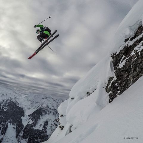 Ein Skifahrer in grün-schwarzer Kleidung springt vor dem Hintergrund eines bewölkten Himmels und schneebedeckter Berge von einer schneebedeckten Klippe. Der Skifahrer befindet sich in der Luft, die Skier zeigen nach vorne.