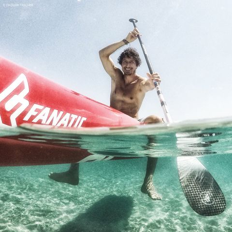 Eine Person paddelt auf einem roten Stand-Up-Paddleboard über klares blaues Wasser. Das Bild wurde halb unter Wasser aufgenommen und zeigt sowohl den Paddler als auch den unter Wasser liegenden Teil des Boards und des Paddels. Die Person scheint zu lächeln und die Aktivität zu genießen.