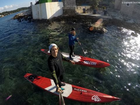 Zwei Personen paddeln auf klarem, blauem Wasser in der Nähe einer felsigen Küste. Sie tragen Neoprenanzüge und benutzen rote Paddleboards. Die Sonne scheint und im Hintergrund sind Gebäude zu sehen. Eine Person hält ein Selfie-Stick und fängt die Szene von oben ein.
