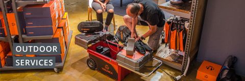 Ein Mann sitzt und arbeitet an einem Paar schwarzer Stiefel neben einem roten Wagen mit Werkzeugen und Kisten. In den Regalen daneben stehen orangefarbene Kisten und hängende Ausrüstung. Auf der linken Seite ist ein Schild mit der Aufschrift „Outdoor Services“ zu sehen.