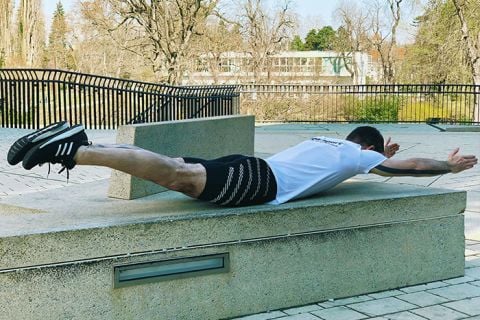 Eine Person in Sportkleidung führt auf einer Betonfläche im Freien einen Superman-Plank aus. Sie liegt auf dem Bauch, hat die Arme nach vorne ausgestreckt und die Beine gerade nach hinten, um eine Flugposition zu simulieren. Im Hintergrund sind Bäume und ein Gebäude zu sehen.