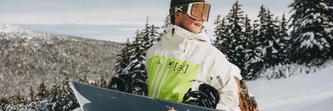 Eine Person in einer grün-weißen Jacke steht mit einem Snowboard in der Hand auf einem schneebedeckten Berghang. Sie trägt eine Schutzbrille und eine Mütze. Im Hintergrund sind schneebedeckte Bäume zu sehen.