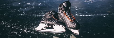 Ein Paar schwarze Hockey-Schlittschuhe mit orangefarbenen Schnürsenkeln steht auf einer Eisbahn im Freien. Die glänzende Eisfläche weist Schlittschuhspuren auf, was darauf schließen lässt, dass sie für Aktivitäten verwendet wurde. Die Schlittschuhe stehen leicht schief, was den ungezwungenen Charakter der Szene verstärkt.