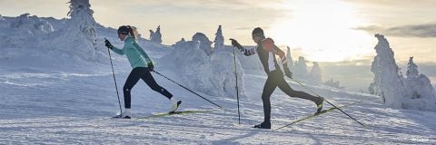 Zwei Personen laufen in einer verschneiten Landschaft unter einem bewölkten Himmel Skilanglauf. Sie tragen Wintersportkleidung und sind von schneebedeckten Bäumen umgeben. Die Sonne steht tief am Horizont und verbreitet einen warmen Schein.