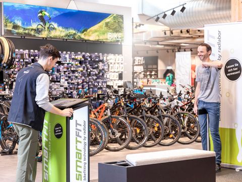 Zwei Personen in einem Fahrradladen beschäftigen sich mit einer Auslage. Einer steht an einem Kiosk, während der andere lächelnd darauf deutet. Hinter ihnen sind Reihen von Fahrrädern und ein großes Fahrradfoto zu sehen. Es gibt helle Beleuchtung und moderne Designelemente.