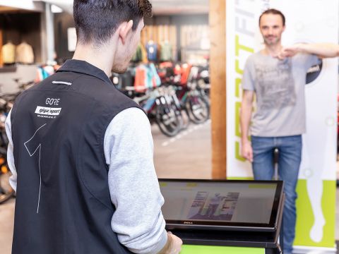 Ein Mitarbeiter in einer GORE-Jacke hilft einem Kunden, der vor einem Anprobebildschirm steht. Der Laden ist hell und modern, im Hintergrund sind Fahrräder und Kleidung ausgestellt.