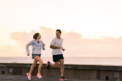 Ein Mann und eine Frau joggen bei Sonnenaufgang Seite an Seite eine Strandpromenade entlang. Der Himmel ist mit weichen Wolken bedeckt, beide tragen Sportkleidung und Turnschuhe, lächeln und schauen sich an.