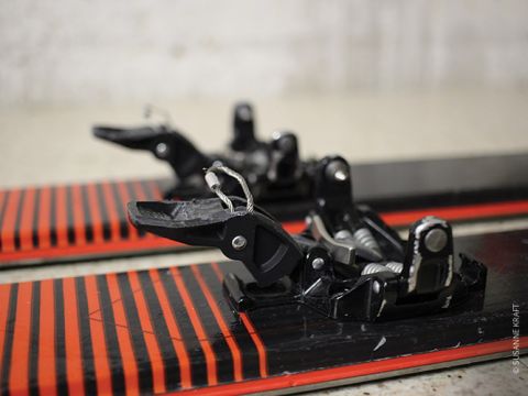 Nahaufnahme von Skibindungen, die an einem Paar Ski montiert sind. Die Ski haben ein schwarz-rot gestreiftes Design. Das Bild konzentriert sich auf den vorderen Teil der Bindungen und zeigt ihre Metall- und Kunststoffkomponenten. Der Hintergrund ist unscharf.