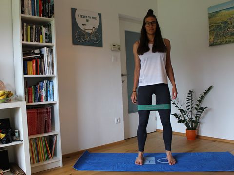Eine Person steht in einem Raum auf einer blauen Trainingsmatte. Sie trägt ein weißes Tanktop und schwarze Leggings und hat ein grünes Widerstandsband um die Oberschenkel. Hinter ihr befinden sich Bücherregale, eine Pflanze und ein Wandbild mit Fahrradmotiv.