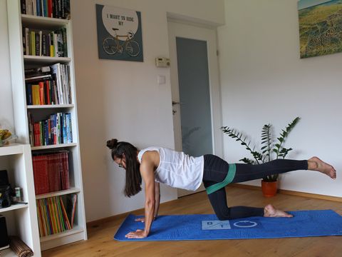 Eine Frau trainiert auf einer blauen Yogamatte mit einem Widerstandsband um ihre Beine. Sie befindet sich in einem Raum mit einem Bücherregal, Wandkunst und einer Topfpflanze. Ihr linkes Bein ist nach hinten ausgestreckt und ihr rechtes Knie liegt auf der Matte.