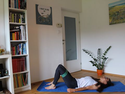 Eine Person liegt auf einer blauen Trainingsmatte in einem Wohnzimmer und führt eine Glute Bridge-Übung mit einem Widerstandsband um die Oberschenkel aus. Im Hintergrund sind ein Bücherregal und eine Topfpflanze zu sehen. An der Wand hängt ein Kunstwerk mit Fahrradmotiv.