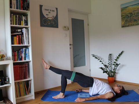 Eine Person führt in einem Raum auf einer blauen Matte eine Glute Bridge-Übung durch. Sie hebt ein Bein an, während das andere gebeugt ist und von einem Widerstandsband gestützt wird. Im Hintergrund sind ein Bücherregal und eine Pflanze zu sehen.