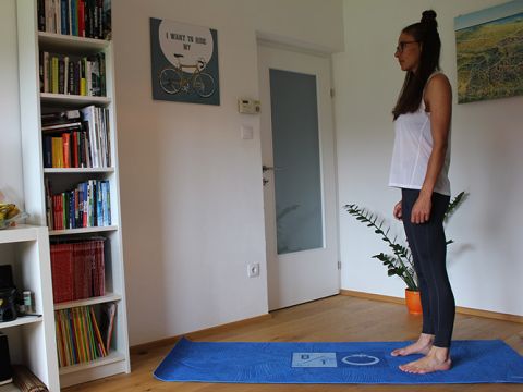 Eine Frau steht auf einer blauen Yogamatte in einem Raum. Sie trägt ein weißes ärmelloses Oberteil und schwarze Leggings. Hinter ihr steht ein Bücherregal mit Büchern, einem Wandposter mit einem Fahrrad, einer Tür und einer Topfpflanze.