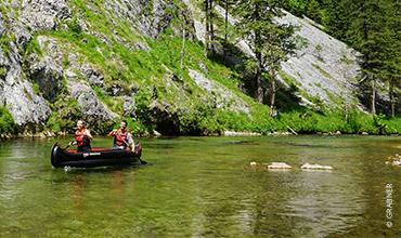 Zwei Personen fahren Kanu auf einem klaren, seichten Fluss, umgeben von üppigem grünem Gras und steilen felsigen Hügeln. Das Wasser ist ruhig und die Sonne scheint, was eine ruhige Atmosphäre schafft. Im Hintergrund sind Bäume zu sehen.