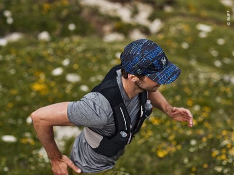 Eine Person läuft im Freien auf einer Wiese. Sie trägt eine blau gemusterte Mütze, ein graues T-Shirt und eine schwarze Trinkweste. Das Gelände ist mit gelben Blumen und Steinen übersät. Der Läufer ist konzentriert und bewegt sich zügig.