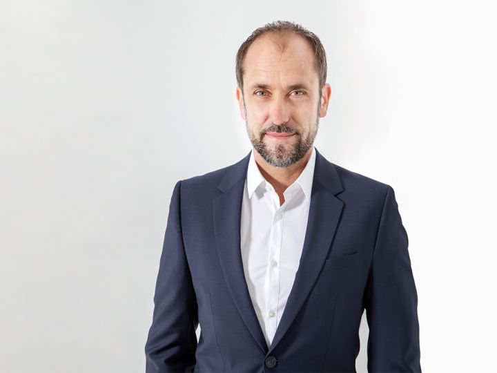 Ein Mann mit kurzen Haaren und Bart in einem marineblauen Anzug und einem weißen Hemd steht vor einem schlichten weißen Hintergrund und blickt mit einem leichten Lächeln in die Kamera.