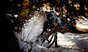 Eine Person in Winterkleidung fährt mit einem Mountainbike durch den Schnee in einem Wald und wirbelt dabei Schnee auf. Sonnenlicht fällt durch die Bäume und hebt die Radfahrszene und die winterliche Umgebung hervor.