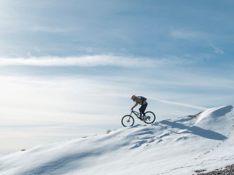 Eine Person mit Helm fährt unter einem klaren blauen Himmel mit dem Fahrrad über einen schneebedeckten Hügel. Im Schnee sind Reifenspuren sichtbar und die Landschaft ist weitläufig und ruhig, mit sanften Schatten, die von der Sonne geworfen werden.