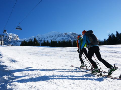 Zwei Skifahrer in Winterkleidung erklimmen mit Skiern und Stöcken einen schneebedeckten Hang. Im Hintergrund sind ein Skilift und ein klarer blauer Himmel zu sehen, und hinter immergrünen Bäumen erheben sich schneebedeckte Berge.