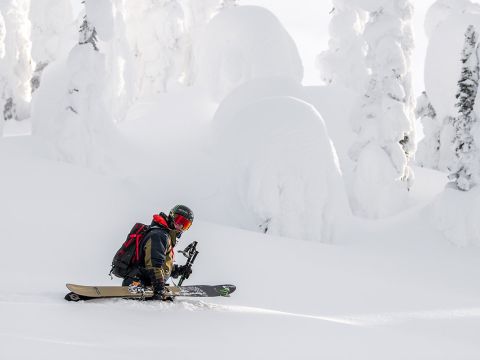 Ein Snowboarder in schwarzer Ausrüstung und rotem Rucksack sitzt mit seinem Snowboard in der Hand auf einem schneebedeckten Hang. Im Hintergrund sind schneebedeckte Bäume im hellen Sonnenlicht zu sehen, die eine ruhige Winterlandschaft schaffen.