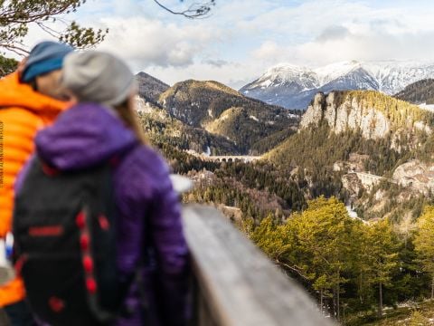 Zwei Menschen in warmen Jacken stehen auf einer Holzplattform und blicken auf eine malerische Berglandschaft. Schneebedeckte Gipfel und dichte Wälder erstrecken sich über die Aussicht, in der Ferne ist ein Steinviadukt zu sehen. Die Szene spielt unter einem hellen, teilweise bewölkten Himmel.