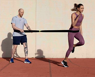 Eine Frau in Sportkleidung läuft nach vorne, während sie an einem Widerstandsband befestigt ist, das ein Mann hinter ihr hält. Sie befinden sich auf einer Laufstrecke mit einer schlichten Wand im Hintergrund. Der Mann trägt ein leichtes Hemd und Shorts, und die Frau trägt einen passenden Sport-BH und Leggings.