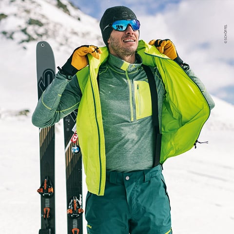 Eine Person mit Sonnenbrille, Mütze und Handschuhen rückt in einer verschneiten Bergregion eine hellgrüne Weste über einer grauen Jacke zurecht. Hinter ihr stehen Skier aufrecht im Schnee.