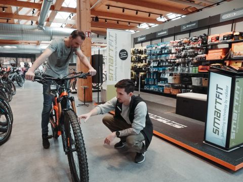 Zwei Personen in einem Fahrradladen: Eine hockt und inspiziert das Hinterrad eines orange-schwarzen Fahrrads, die andere steht neben dem Rad. Im Hintergrund sind Regale mit Fahrradzubehör zu sehen.