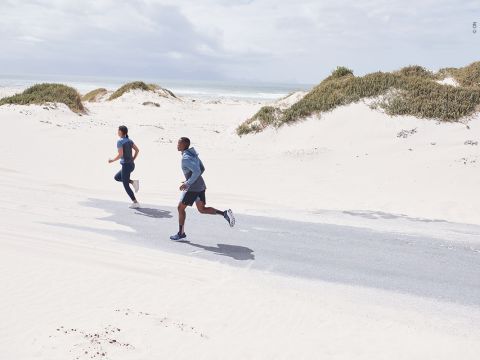 Zwei Personen laufen auf einer asphaltierten Straße durch eine sandige Strandlandschaft. Sie tragen Sportkleidung und der Himmel ist teilweise bewölkt. Um sie herum sind Sanddünen und spärliche Vegetation zu sehen.