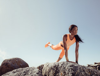 Eine Frau in leuchtend orangefarbener Sportkleidung balanciert auf Felsen und macht dabei eine Plankenübung, wobei sie ein Bein nach hinten streckt. Der Himmel ist klar und blau, was auf einen sonnigen Tag schließen lässt.