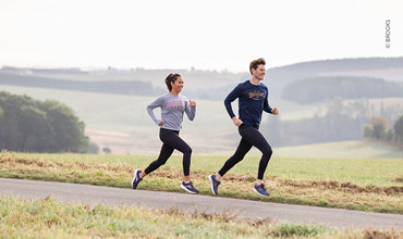 Eine Frau und ein Mann joggen auf einem gepflasterten Weg durch eine malerische Landschaft. Der Himmel ist bedeckt, im Hintergrund sind Grasfelder mit entfernten Hügeln zu sehen. Beide tragen Sportkleidung.