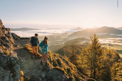 Zwei Wanderer, ein Mann und eine Frau, gehen einen felsigen Pfad entlang, von dem aus man ein Tal überblicken kann. Die Sonne geht hinter den Bergen in der Ferne unter und wirft einen warmen Schein auf eine Landschaft mit sanften Hügeln und vereinzelten Wolken. Unten sind Bäume und grüne Felder zu sehen.