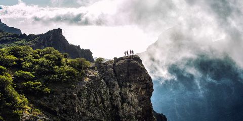 Eine Gruppe von Wanderern steht auf einer Felsklippe, deren Silhouette sich vor einem wolkigen Himmel abzeichnet. Die Landschaft zeichnet sich durch dramatische Felsformationen und viel Grün aus, im Hintergrund sind neblige Berge zu sehen.