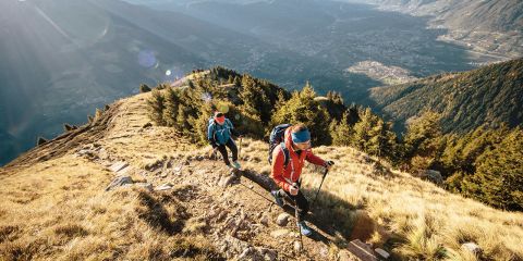 Zwei Wanderer steigen mit Trekkingstöcken einen grasbewachsenen Bergpfad hinauf. Sonnenlicht durchflutet das weite Tal und die Berge in der Ferne. Beide tragen Rucksäcke und Outdoor-Ausrüstung und haben eine malerische Aussicht auf zerklüftetes Gelände und dichte Bäume.