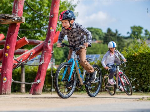 Ein Kind mit einem roten Helm fährt energisch auf einem blauen Fahrrad auf einem Feldweg, im Hintergrund ist ein Spielplatz zu sehen. Ein Erwachsener folgt ihm auf einem roten Fahrrad und trägt einen weißen Helm. Die Szene ist hell und sonnig.