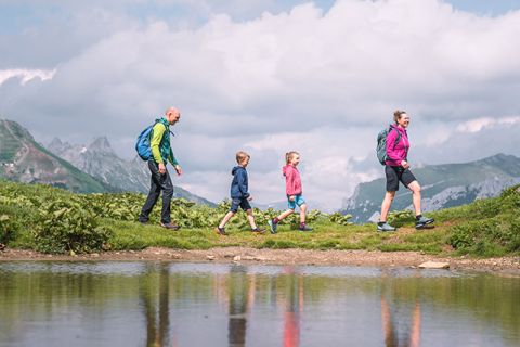 Eine vierköpfige Familie wandert auf einem malerischen, grasbewachsenen Pfad mit einem ruhigen Teich im Vordergrund. Im Hintergrund sind Berge unter einem teilweise bewölkten Himmel zu sehen. Jedes Mitglied trägt farbenfrohe Wanderkleidung und einen Rucksack.